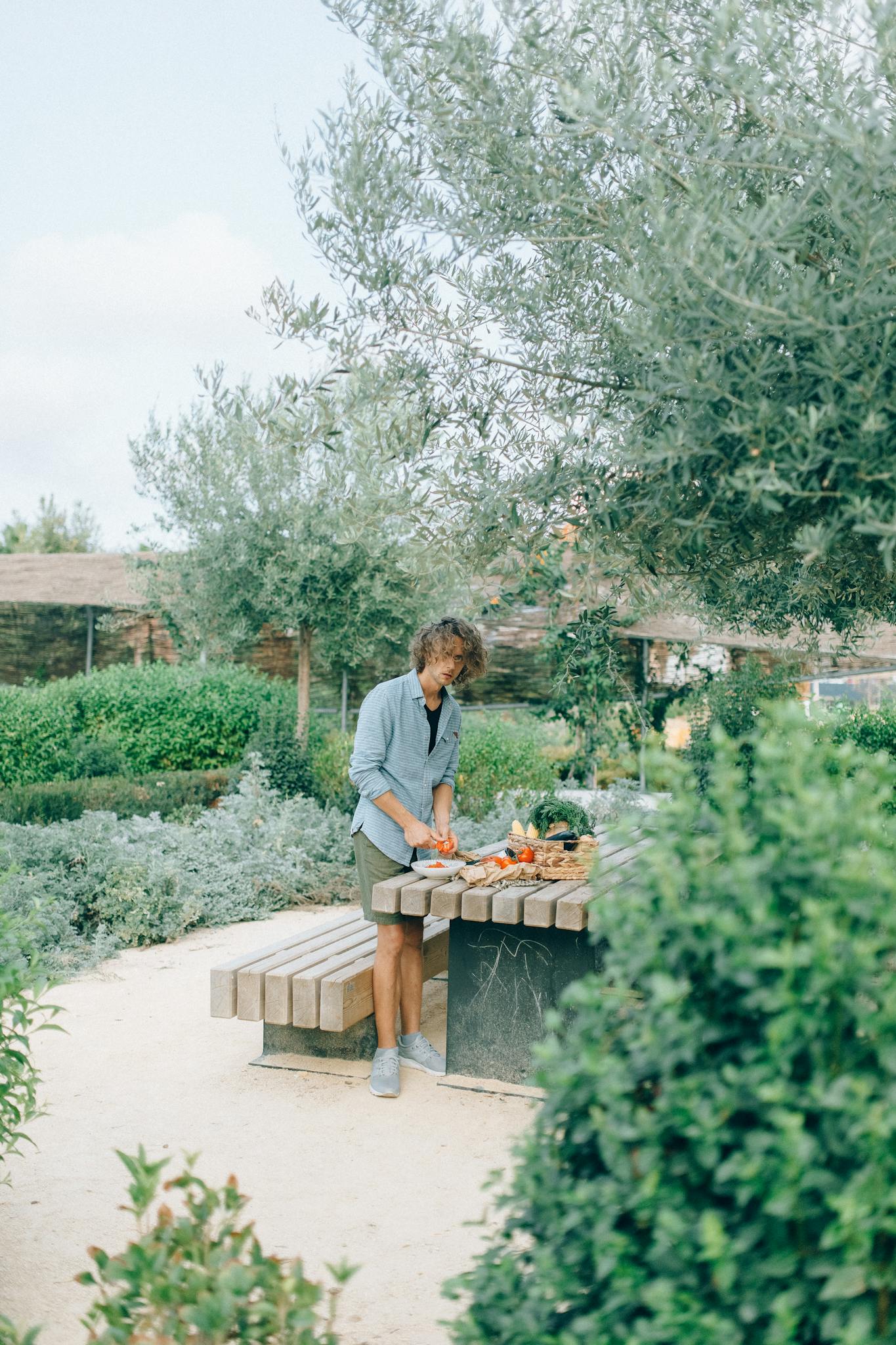 A man cuts vegetables on a wooden table in a lush outdoor garden setting.