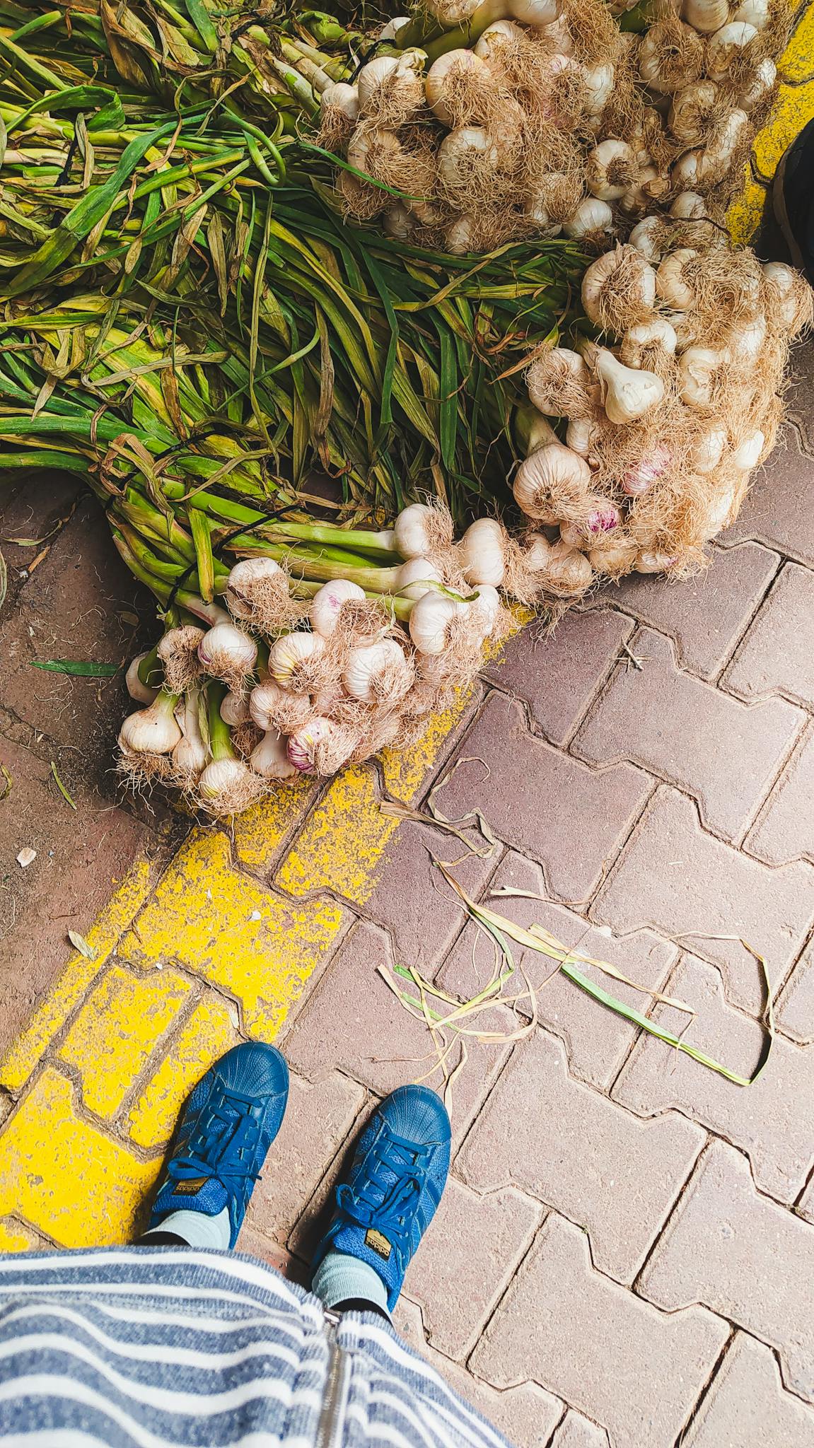 Fresh bunches of garlic on a market pavement next to a person's blue shoes.